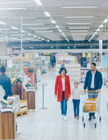 At the Supermarket: Happy Family of Three, Holding Hands, Walks Through Fresh Produce Section of the Store. Father, Mother and Daughter Having Fun Time Shopping. High Angle Panoramic Shot.